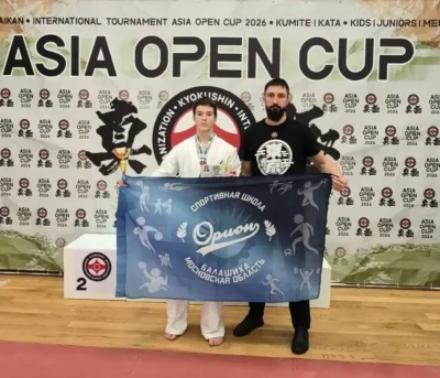 Спортсмен с медалью и представитель команды с флагом спортивной школы «ОРИОН» на фоне баннера ASIA OPEN CUP 2026.